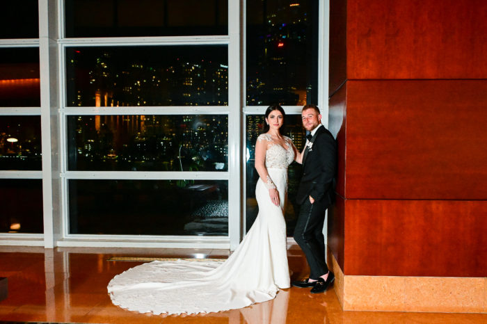 A bride and groom pose for the camera in front of a window.
