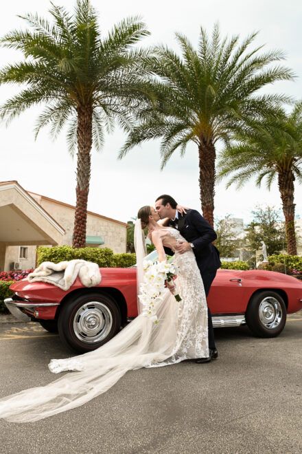 A bride and groom kissing in front of a red car.
