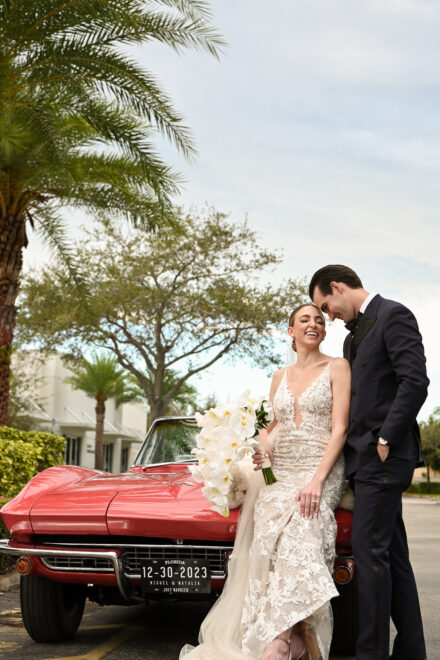 A bride and groom standing next to a red car.