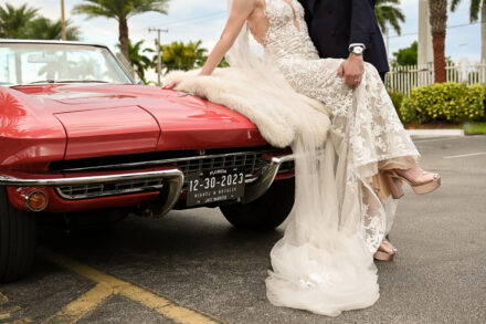 A man and woman in wedding dress next to red car.